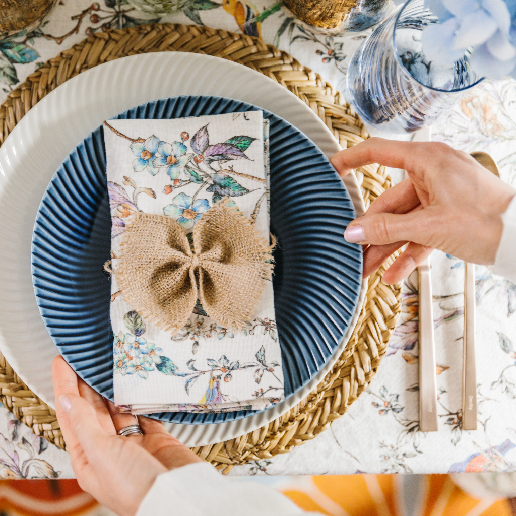 Top down view of hands placing navy arc pattern start plate on top of white arc pattern dinner plate from Denby Porcelain range. The crockery is set with Wildflower linen napkins, hessian napkin bows and a natural seagrass placemat.