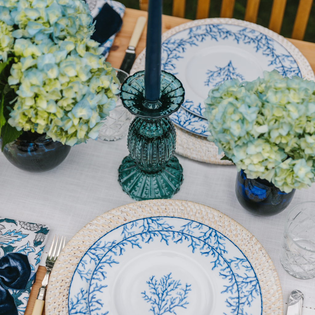 Top down view of scalloped blue glass candle holder set on white cotton table runner next to blue coral plate sets