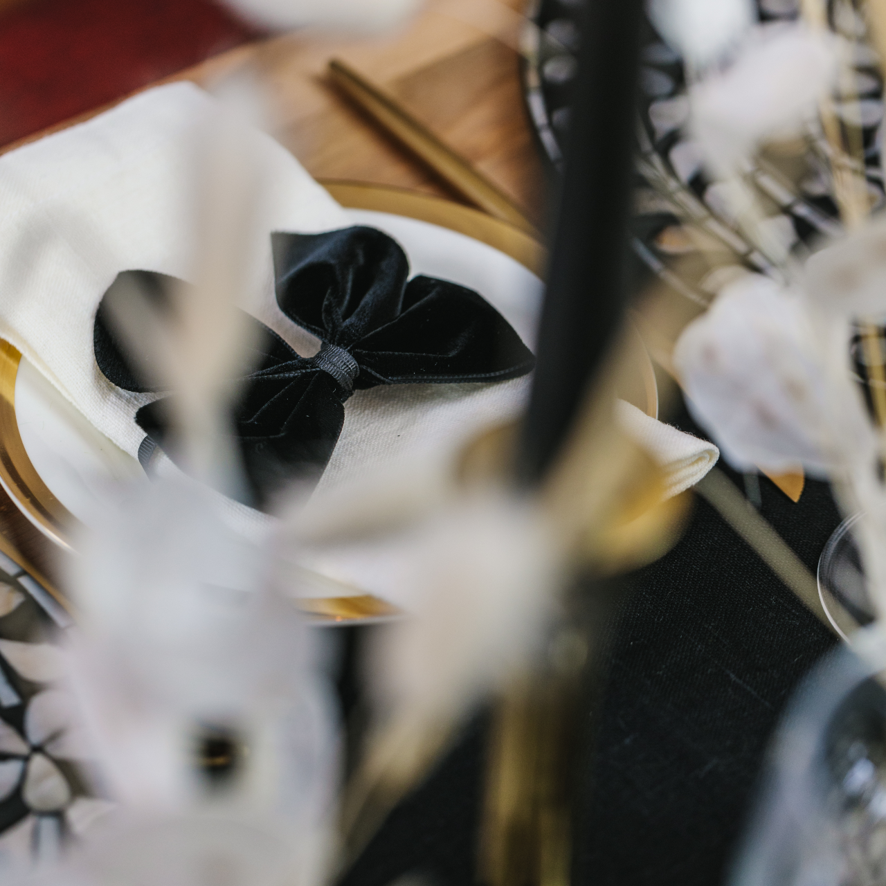 Peering through dried grasses and wheats to a black tapered candle and black velvet napkin bows set on top of 100% white linen napkins all part of Magical Monochrome tablescape