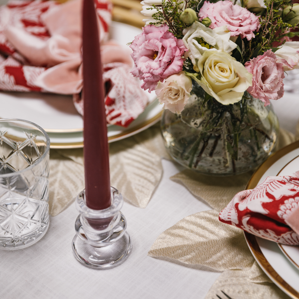 Deep red taper candle set in a short tulip shaped glass candle holder, clear glass bud vase with posy of pink spring flowers