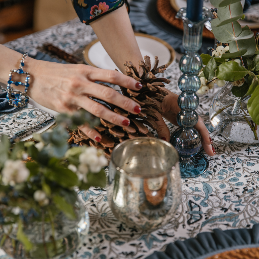 Image of hand placing large pine cone next to powder blue glass candlestick and copper metallic tealight holder on blue block printed tablelcloth.