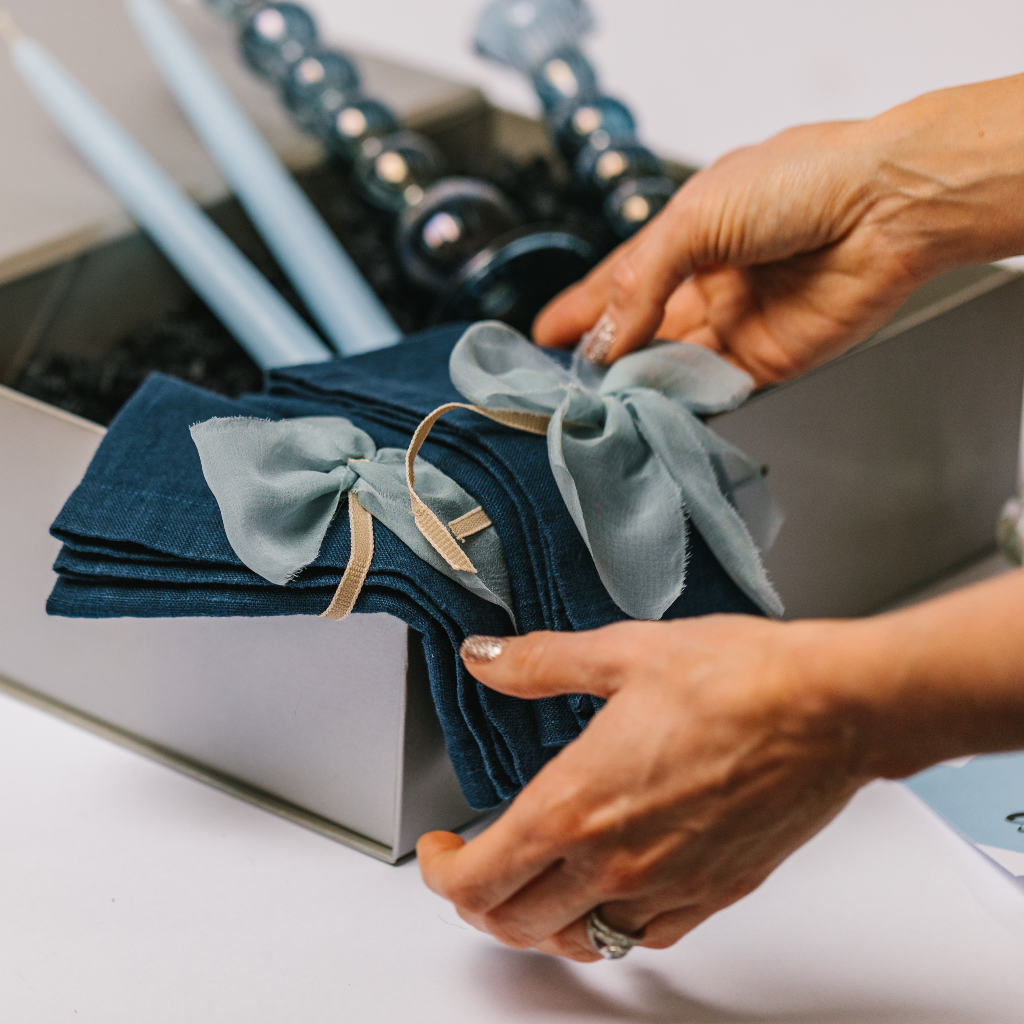 Hands laying out navy linen table napkins with pale blue silk napkin bows. In the background there are hand blown blue glass candle holders and pale blue tapered dinner candles