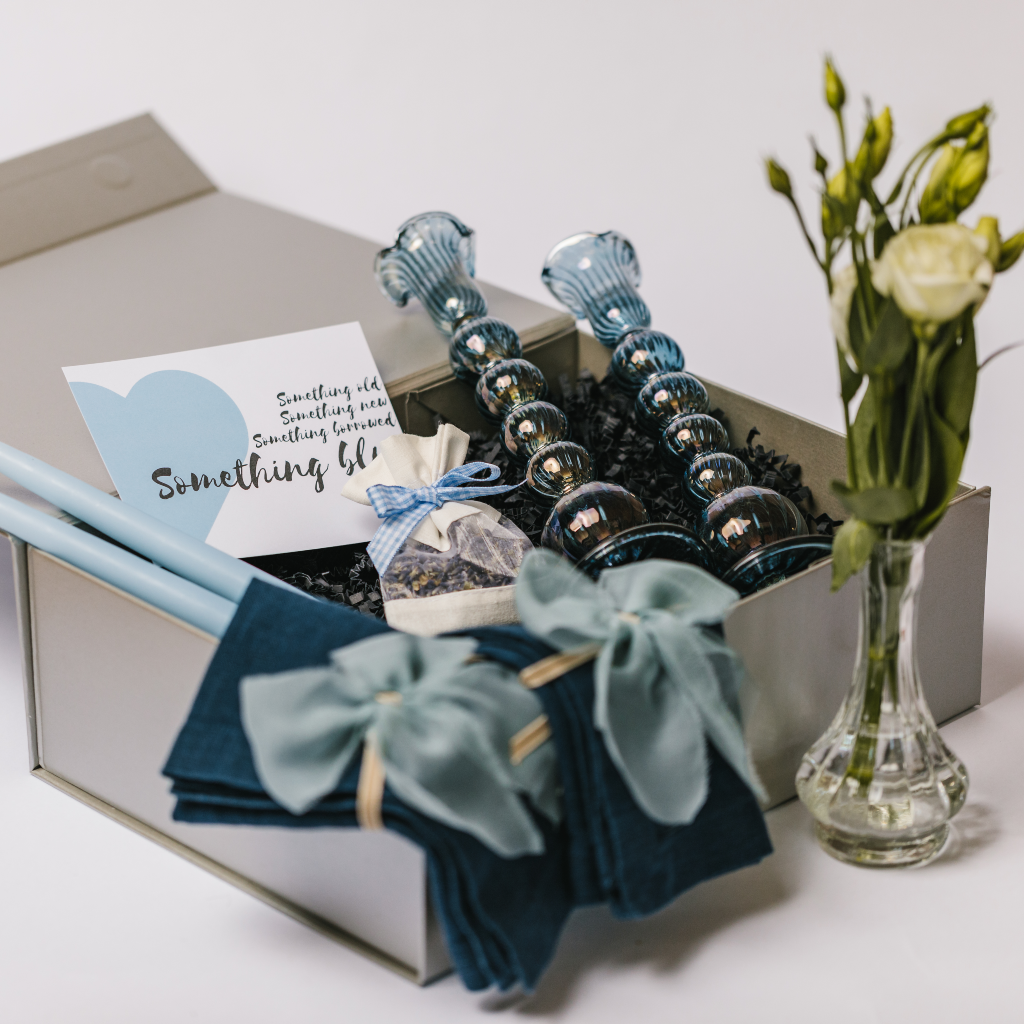 Luxury 'Into the Blue' wedding, anniversary or engagement gift box laid out to show hand blown blue glass candlesticks, a petite bud vases with white flowers display, 100% linen navy napkins, raw edge silk napkin bows and a posy of dried lavender with pale blue gingham tie. The box also contains a 'Something Blue' gift card