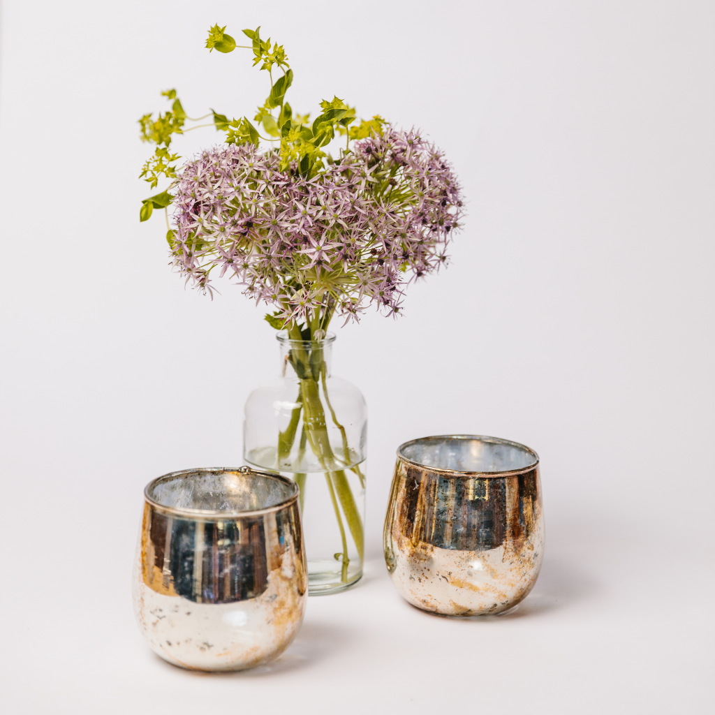Two copper tealight holders set out with an apothecary-style bud bottle and a posy of purple and green flowers