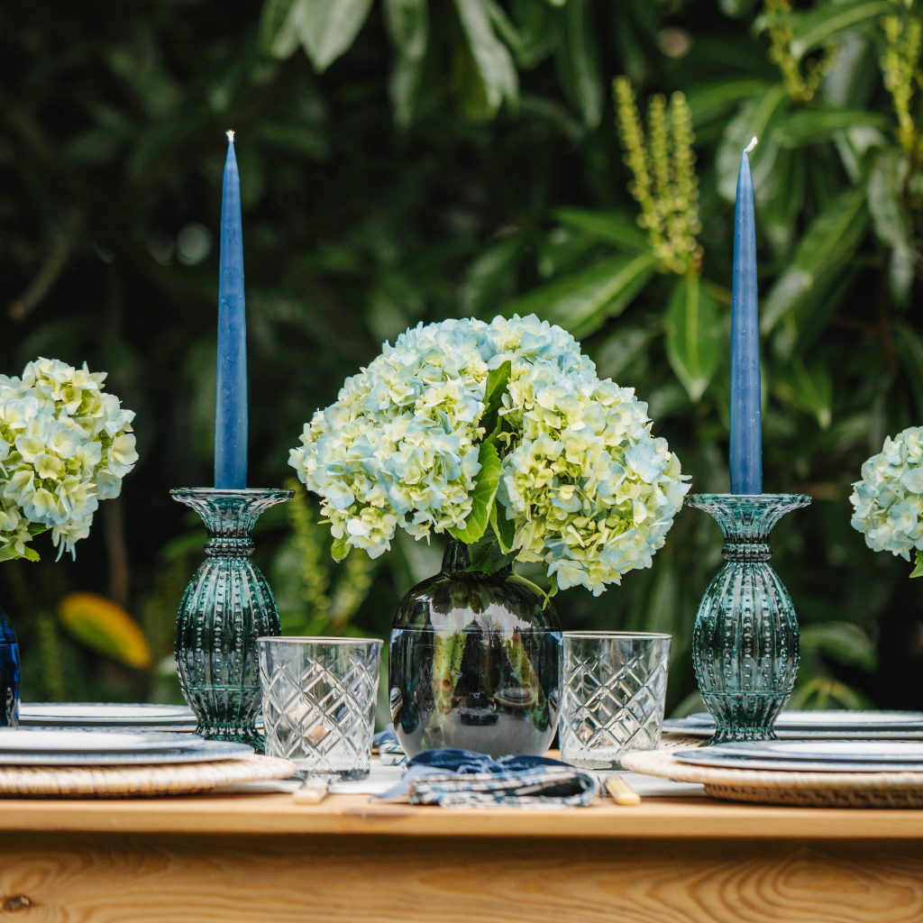 Duo of aquamarine glass candlesticks holding navy tapered candles next to blue dappled glass vase containing hydrangea