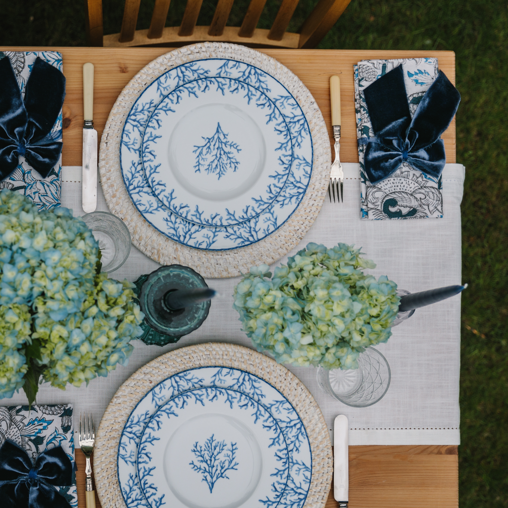 Top down view of two person Santorini blue coral plate set with white cotton table runner, patterned blue and white cotton napkins, navy blue velvet napkin bows and blue hydrangea