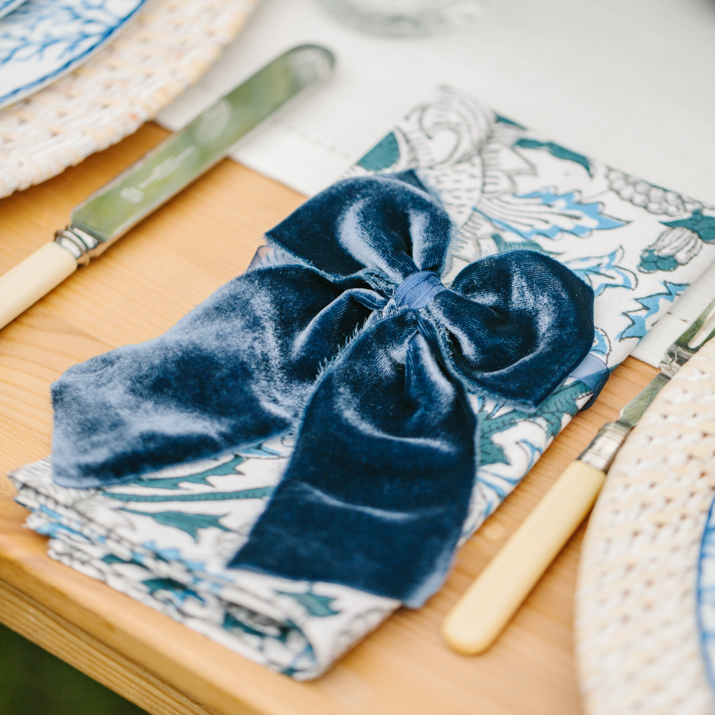 Blue and white Santorini patterned napkin folded and adored with navy blue velvet napkin bow next to vintage cream cutlery and white rattan charger plate