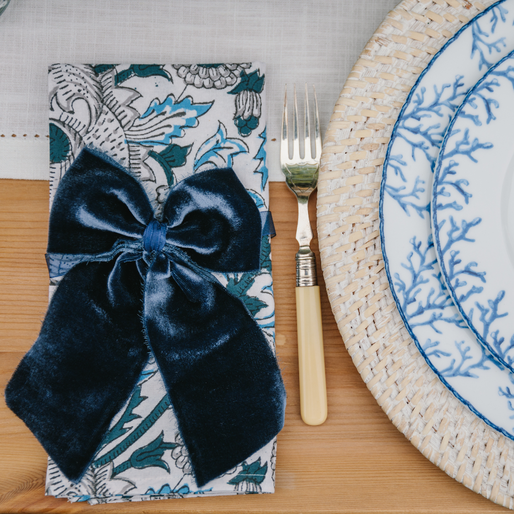 Blue and white Santorini patterned napkin folded and adored with navy blue velvet napkin bow next to vintage cream cutlery, white rattan charger plate and blue coral designed dinner and starter plates