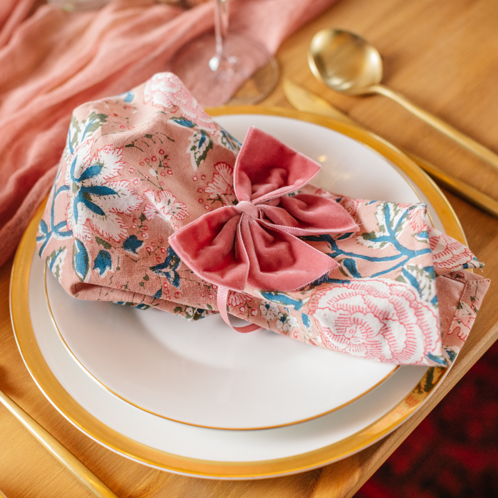 Pale pink floral print cotton napkins ruffled and placed on top of gold trimmed plate and tied with large pink velvet napkin bow. The place setting includes gold cutlery and a pink ruffled cheesecloth table runner.