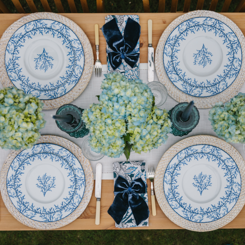 Top down image of Santorini tablescape with blue and white porcelain coral dinner and starter plate sets, white cotton table runner, blue glass candle holders and luxury navy napkin bows
