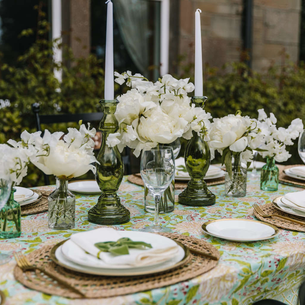 Detailed view of Mint & Moss tableware with statement bottle green glass candle holders, white tapered dinner candles, oval seagrass placemats, gold rimmed side plates and white linen napkins