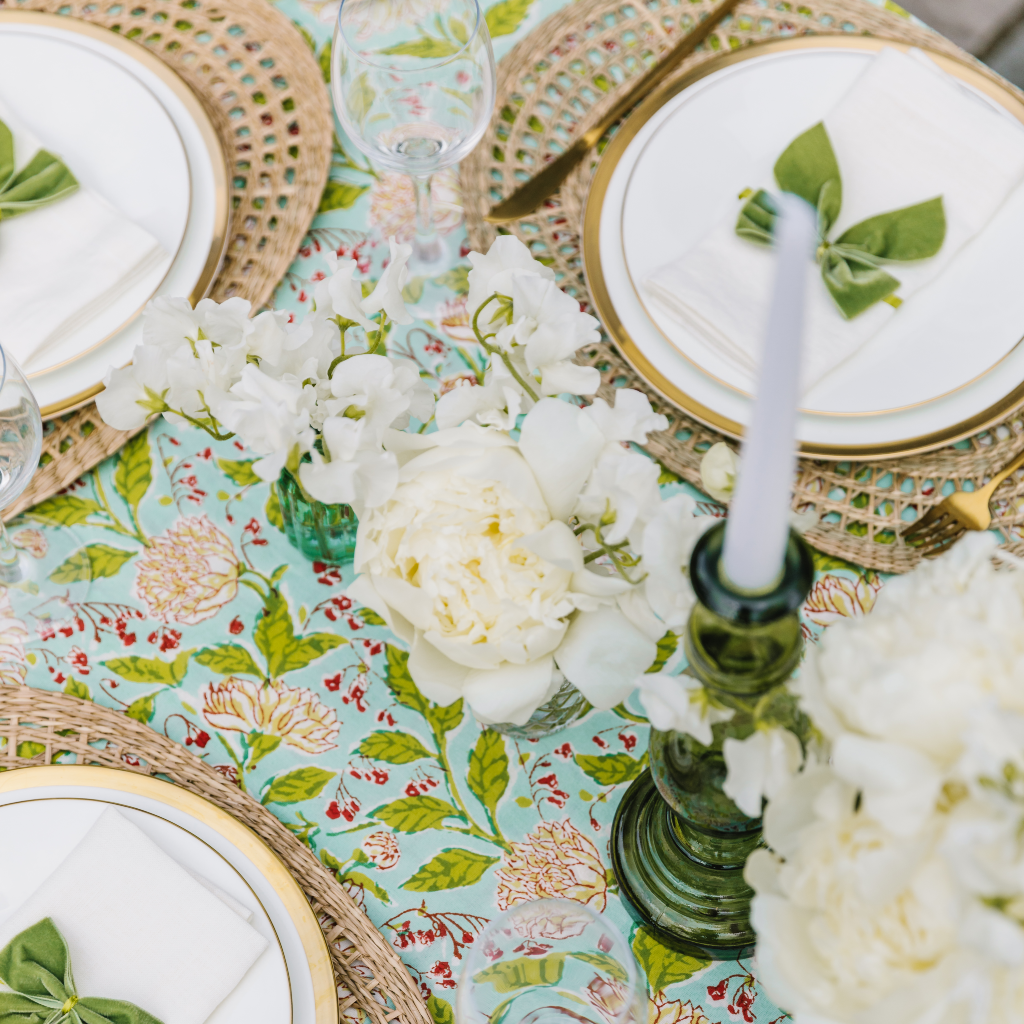 Birds eye view of mint & moss tablecloth including moss green leaf detals, cream flowers and red accents. Place settings include oval seagrass placemats, white linen napkins and green velvet napkin bows