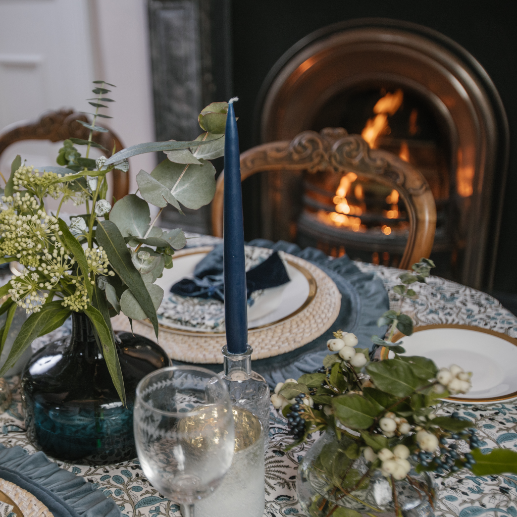 Navy tapered dinner candle in cut glass bottle holder next to navy vase with greenery. The tablescape is set with blue velvet ruffle placemats, navy blue napkin bows and Indian block print table linens.