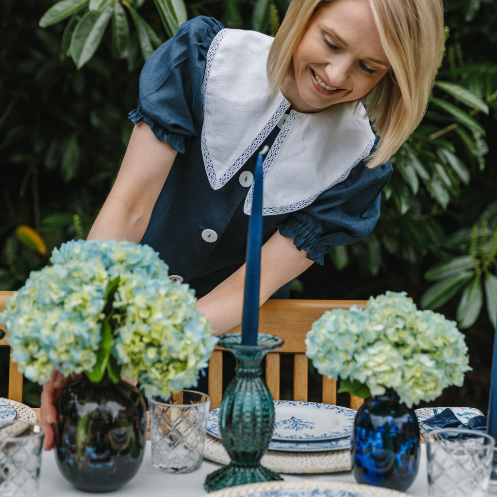 Kate Fairlie, founder of Truffle Tablescapes, setting a blue dappled glass vase onto the Santorini coral tablescape next to an aquamarine cut glass candle holder