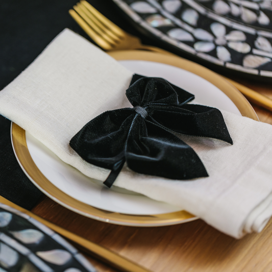 Luxurious black velvet napkin bow sitting on top of 100% white linen napkin and gold trimmed side plate. Black and white mother of pearl inlay charger plates in floral design next to crockery