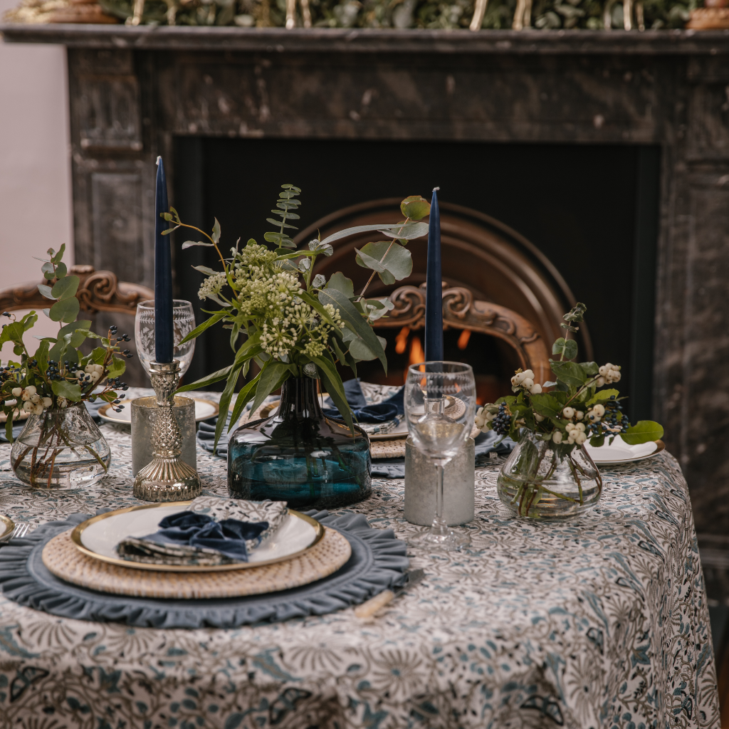 Side of view of Twilight tableware collection set on table in front of black marble fireplace. The tablescape includes blue Indian block print cotton tablecloth and napkins, bourbon blue velvet ruffled placemats, a large navy vase and navy blue tapered dinner candles.