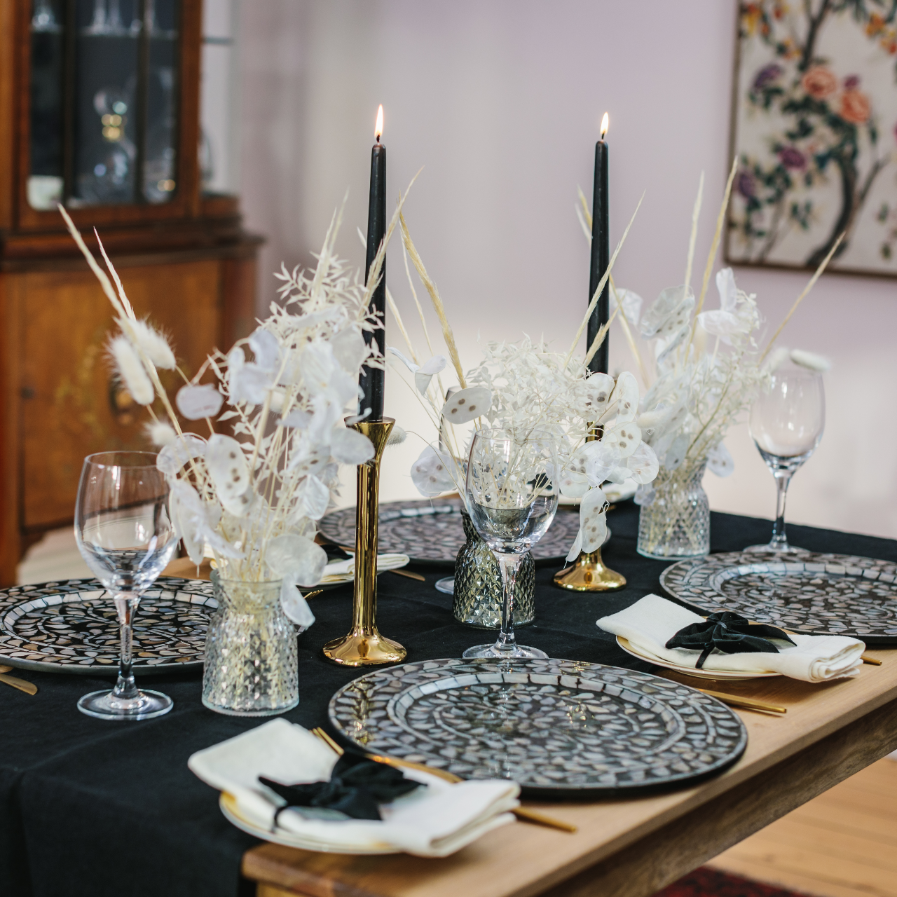 Side view of monochrome tablescape in a box table décor collection including black and white mother of pearl charger plates, sleek brass candlesticks, black tapered dinner candles, cut glass bud vases and dried flower displays with bunny tail and grasses