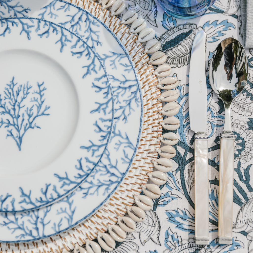Blue and white coral design Santorini starter plate and main plate layered on a rattan and shell-trimmed placemat next to white pearl cutlery