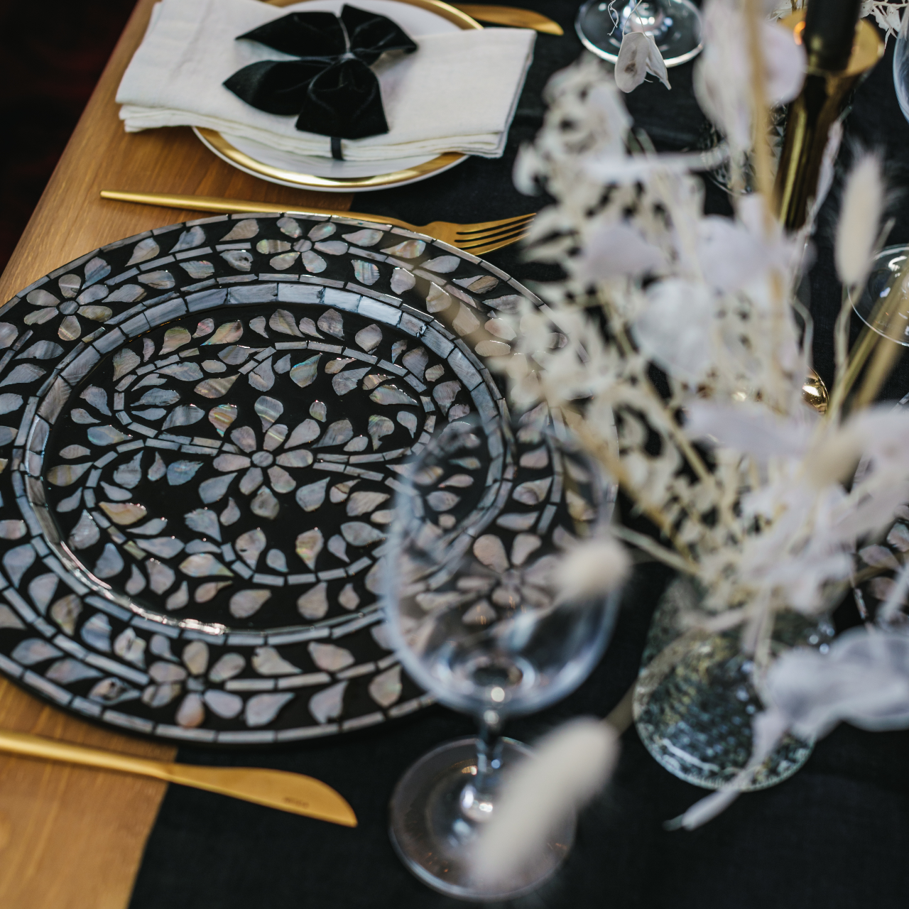 Side image of statement black and white mother of pearl inlay charger plates set with gold cutlery, 100% black linen table runner, white linen napkins, luxe black velvet napkin bows and dried grass posies
