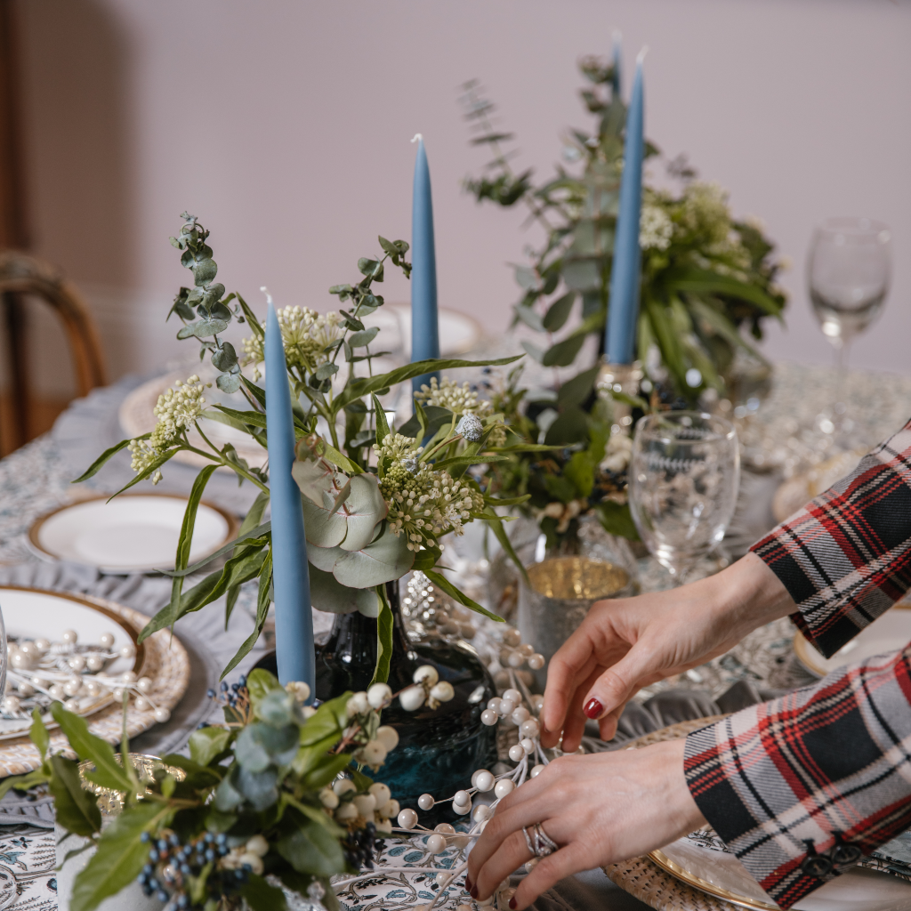 Grey blue tapered candles displayed down the middle of a tablescape with dark blue vase and silver candle holders. Hands can be seen laying a pearl garland on top of an Indian block printed blue tablecloth.