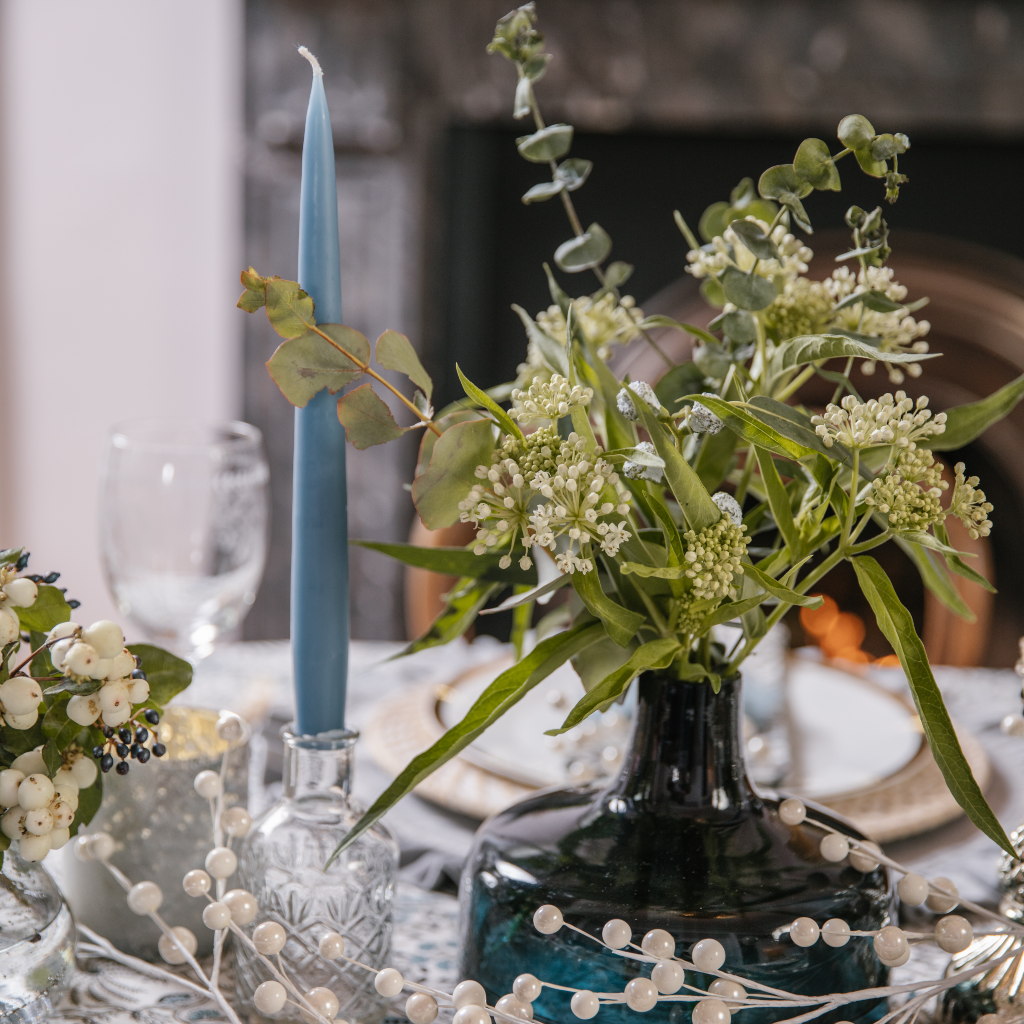 Grey blue tapered dinner candle in cut glass bud bottle situated next to a navy vase containing greenery and white buds.