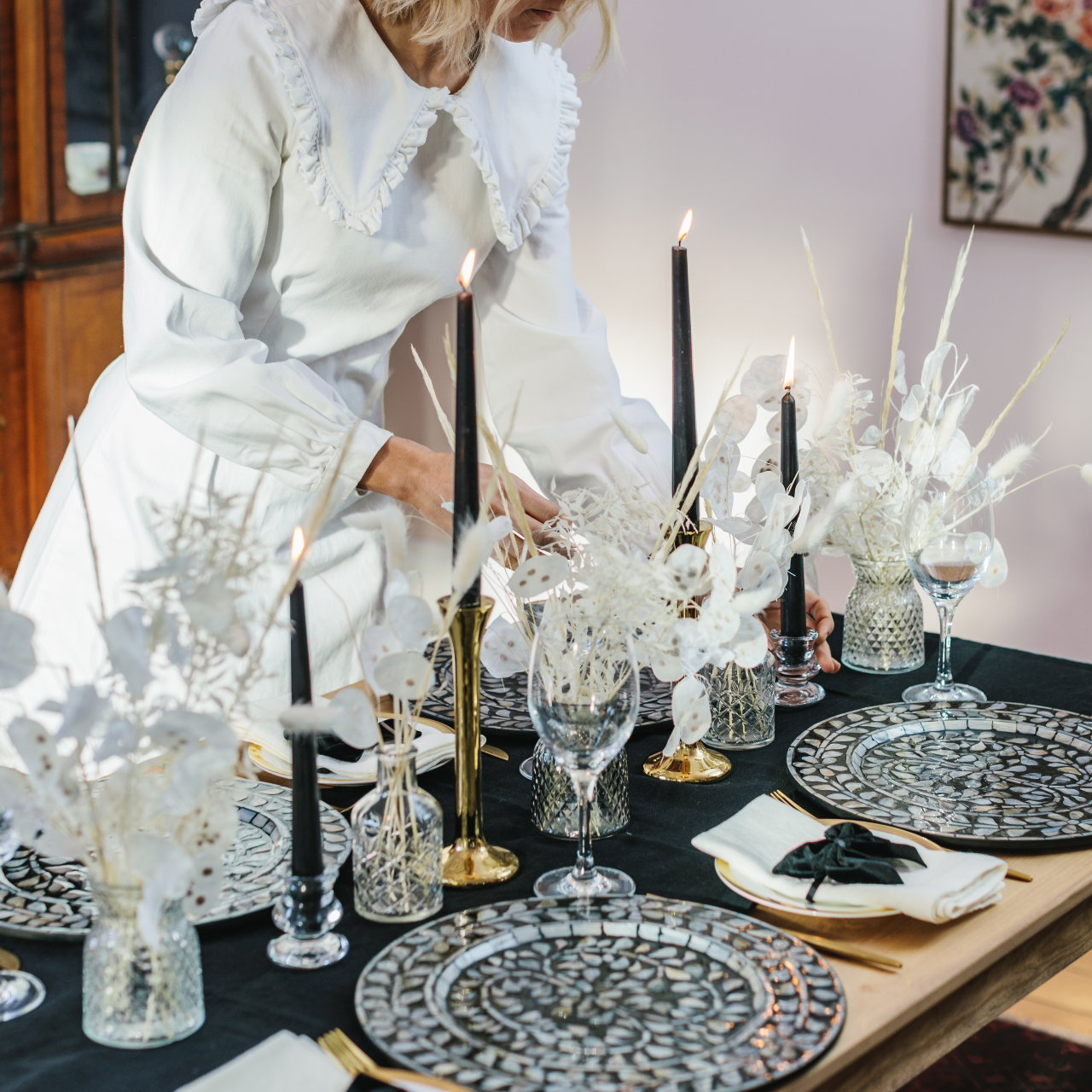 Side view of monochrome tablescape in a box table décor collection including black and white mother of pearl charger plates, sleek brass candlesticks, black tapered dinner candles, cut glass bud vases and dried flower displays with bunny tail and grasses