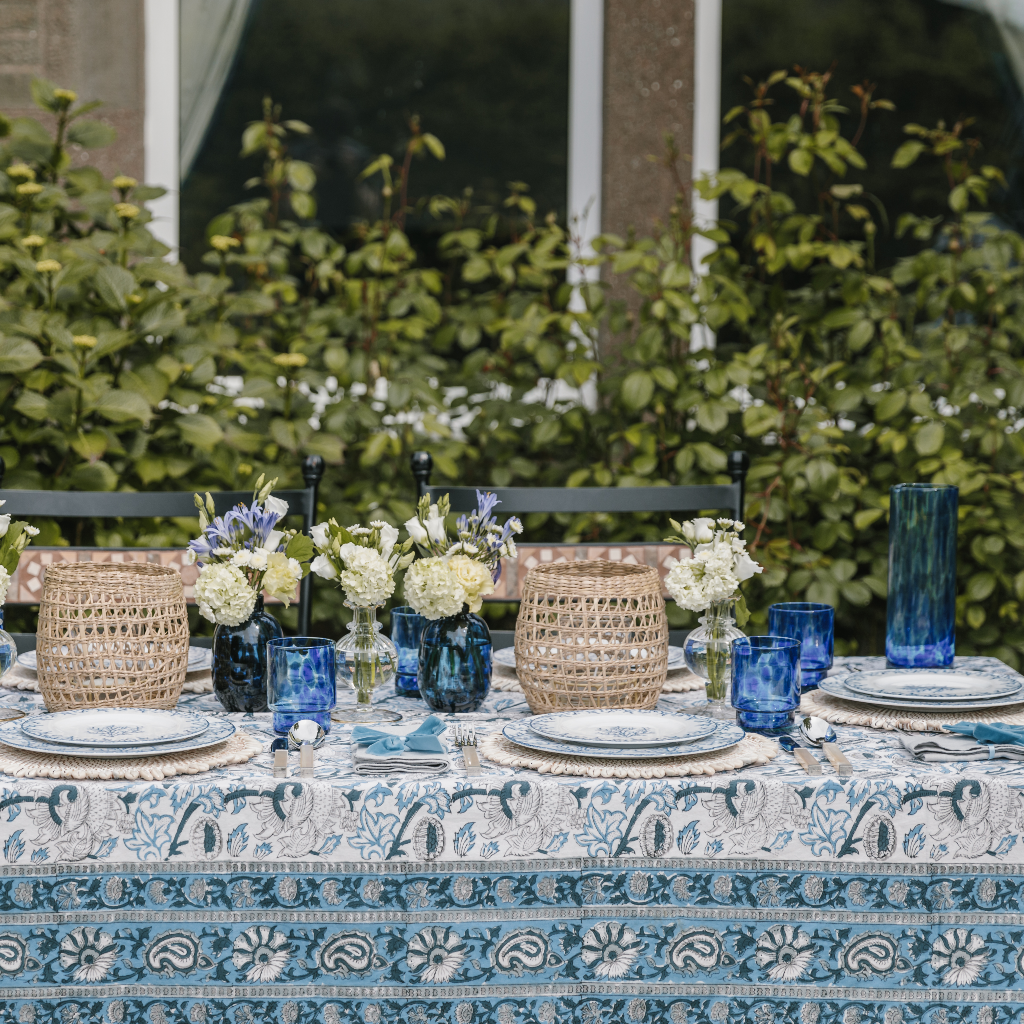The Lapis Blue Tablecloth with navy, white, blue and grey block print pattern set on an al fresco tablescape with blue dappled tumblers and woven seagrass lanterns