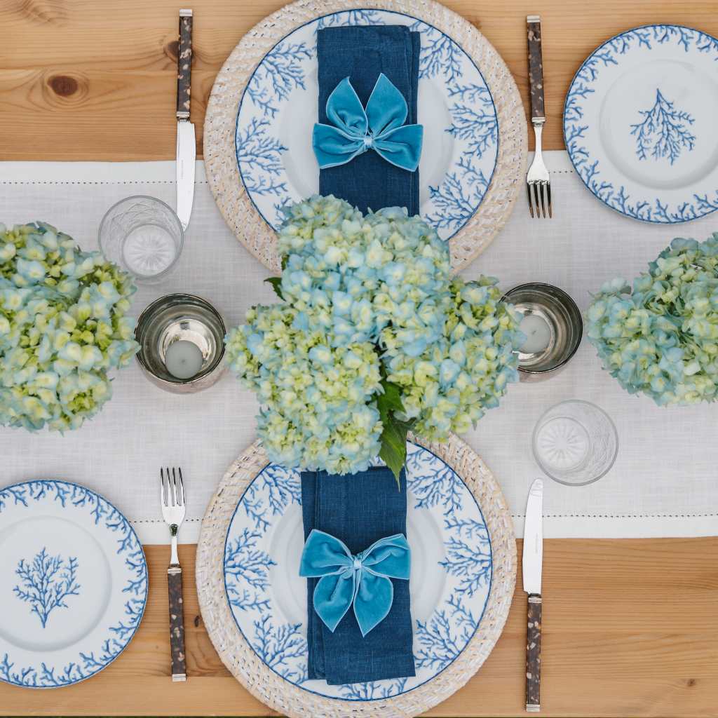 Porcelain cotton table runner with pintuck edging seat with white rattan charger plates, Santorini coral plates, navy linen napkins and kingfisher blue velvet napkin bows