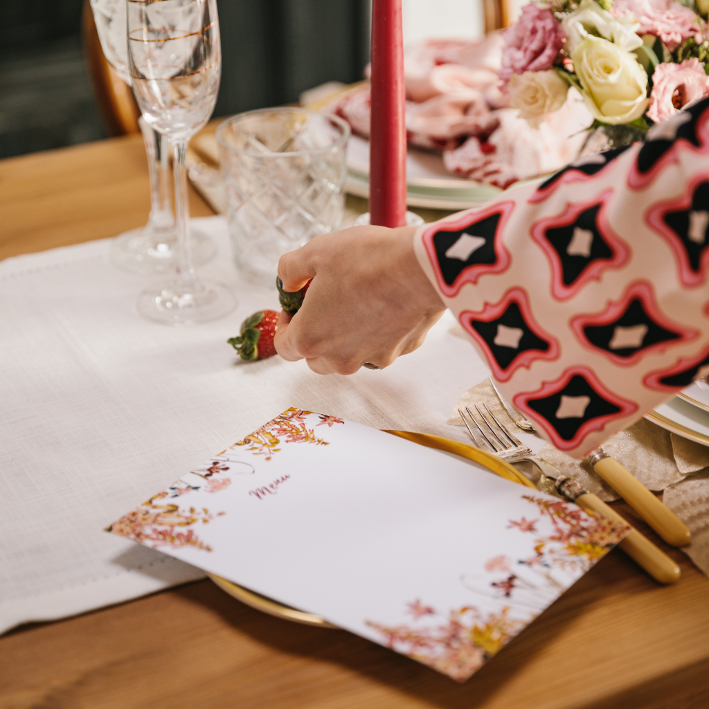 Hands laying strawberries on a white cotton table runner next to a blank bespoke menu card with pink floral deisgn