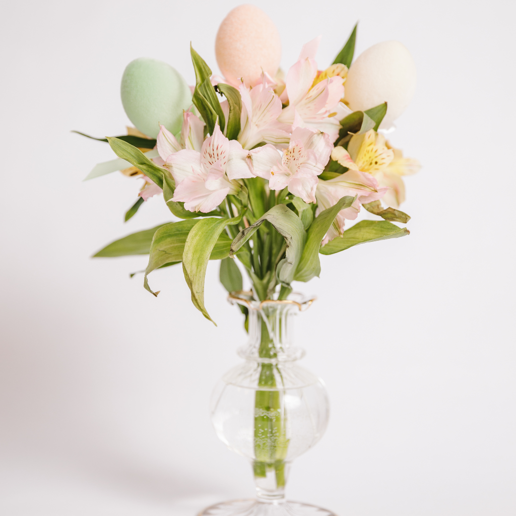 Posy of spring flowers in a ruffled hand blown clear glass bud vase displayed with flocked cream, pink and mint Easter eggs