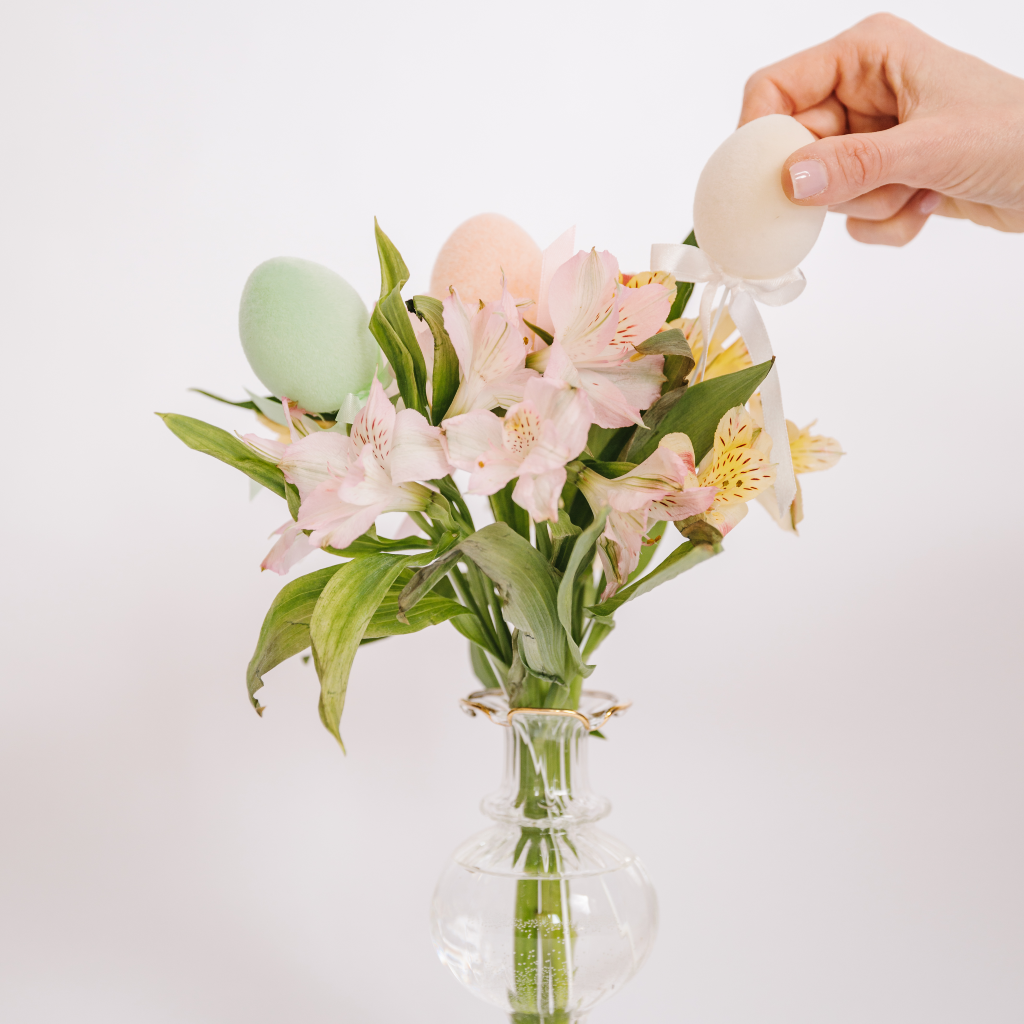 Hand placing a cream flocked Easter egg decoration with ribbon into a posy of pink and green spring flowers displayed in a ruffled gold edged Egyptian bud vase
