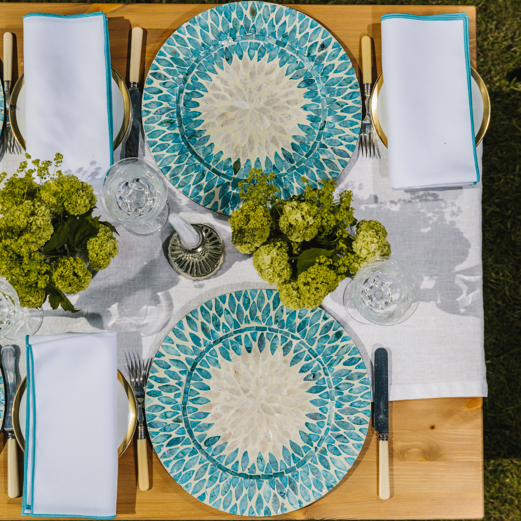 100% white linen tablecloth set with luxury aqua and white mother of pearl charger plates and embroidered trim cotton napkins. Lush green blooms fill the candle of the table along with an ornate silver candle holder and white tapered dinner candle