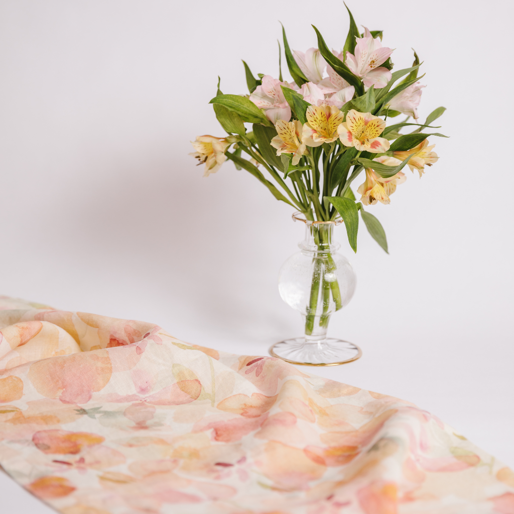 A peach, pink, cream and pale green watercolour pattern dappled table runner draped next to a gold trimmed hand blown ruffled bud vase containing a posy of pink and yellow flowers