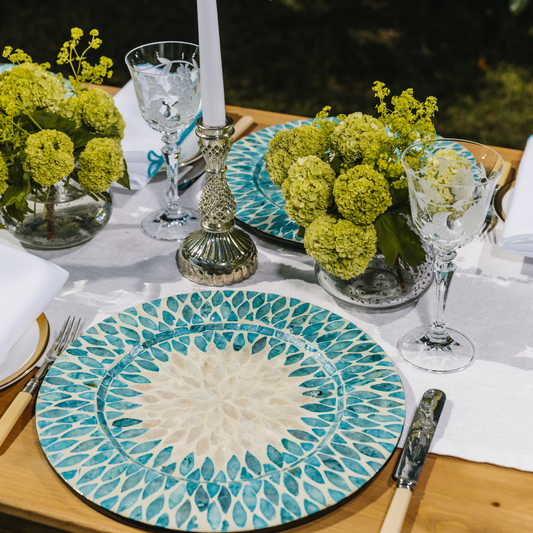 White linen table runner set with fresh green flowers nestled into clear glass bud vases. Aqua mother of pearl charger plates are set with vintage cream handled cutlery and etched glassware