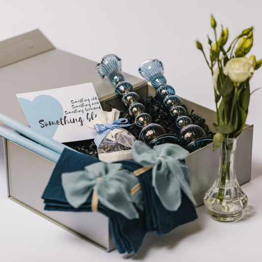 Luxury 'Into the Blue' wedding, anniversary or engagement gift box laid out to show hand blown blue glass candlesticks, a petite bud vases with white flowers display, 100% linen navy napkins, raw edge silk napkin bows and a posy of dried lavender with pale blue gingham tie. The box also contains a 'Something Blue' gift card