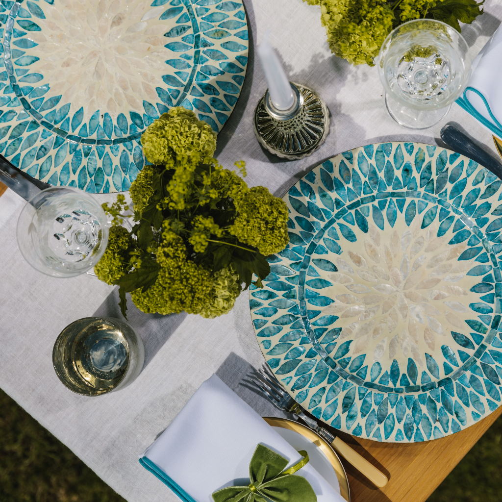 Top down image of white linen table runner set with aqua mother of pearl charger plates, white embroidered trim napkins and luxury moss green velvet napkin bows