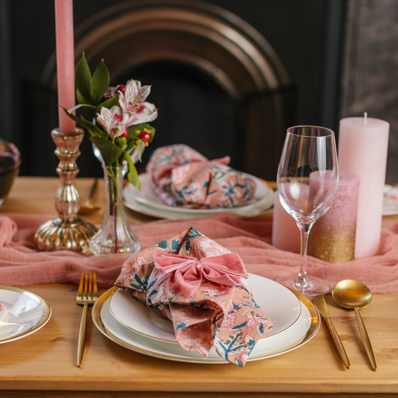 View of pink tablescape set for two people including pink patterned napkins, pink velvet napkin bows, gold cutlery, pink pillar candle cluster, a bud vase and metallic candle holder with pink dinner candle.