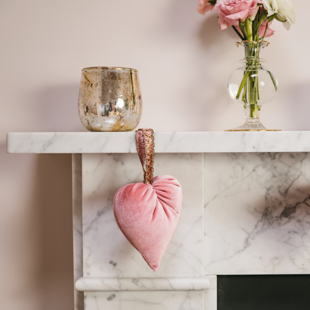 Copper burnished tealight holder on a white marble mantlepiece next to a hand blown clear bud vase with ruffled gold trim. A plush pink velvet heart is draped over the fireplace.