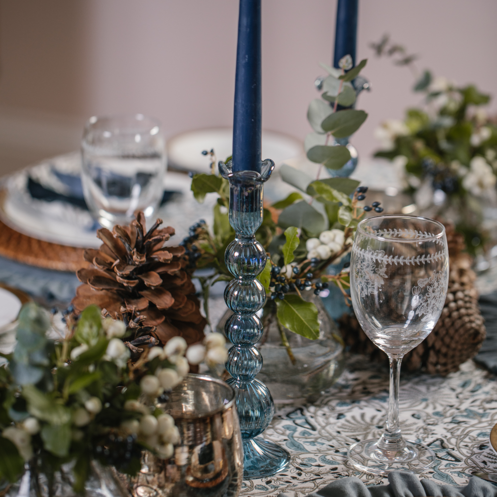 Blue glass tall candle holders with navy tapered candles sea on Truffle's Twilight Tablescape including giant pinecones, copper candle holders, a block printed blue tablecloth and ruffled placemats.