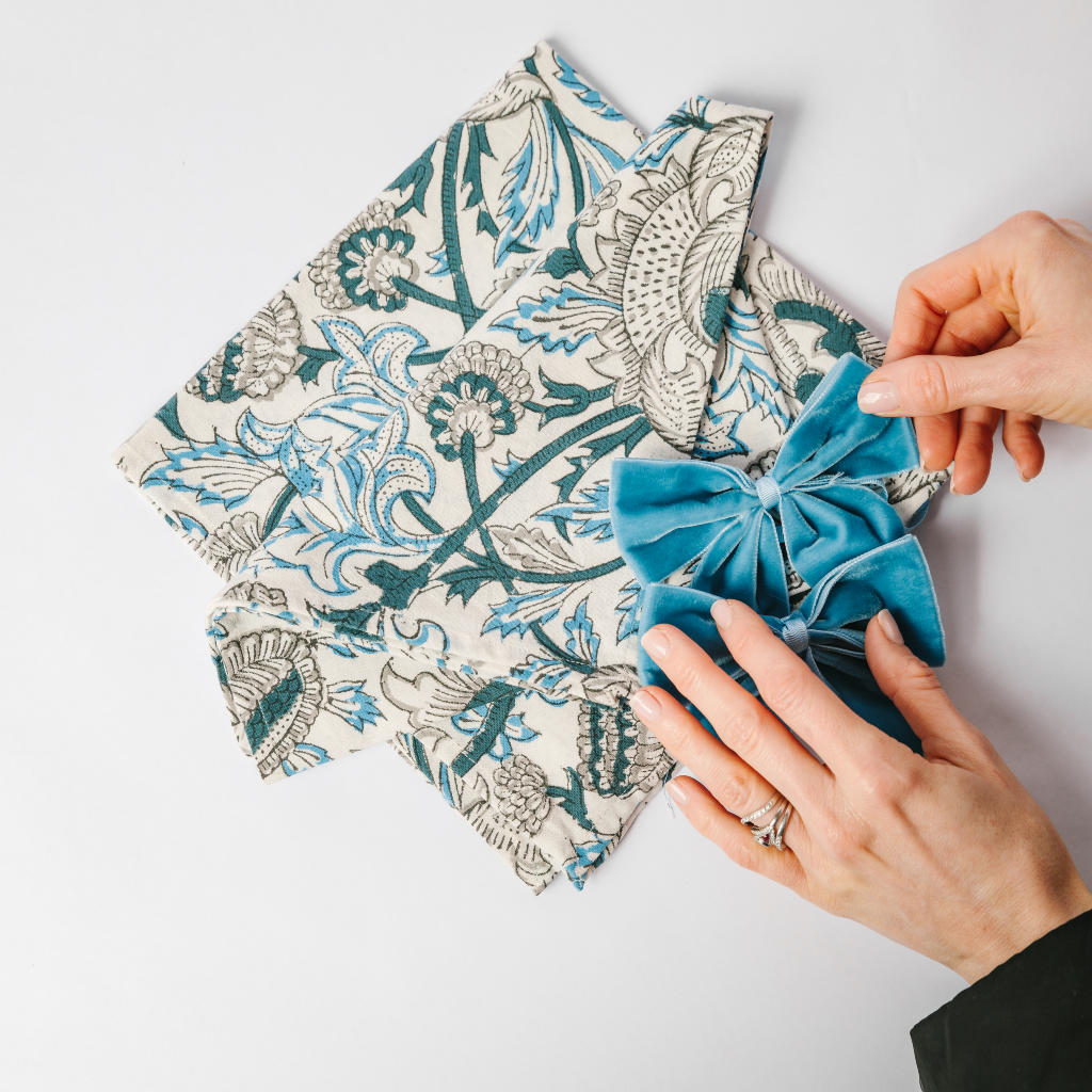 Navy blue, light blue and white block printed pattern cotton napkin folded next to hand reaching out to kingfisher blue velvet napkin bows