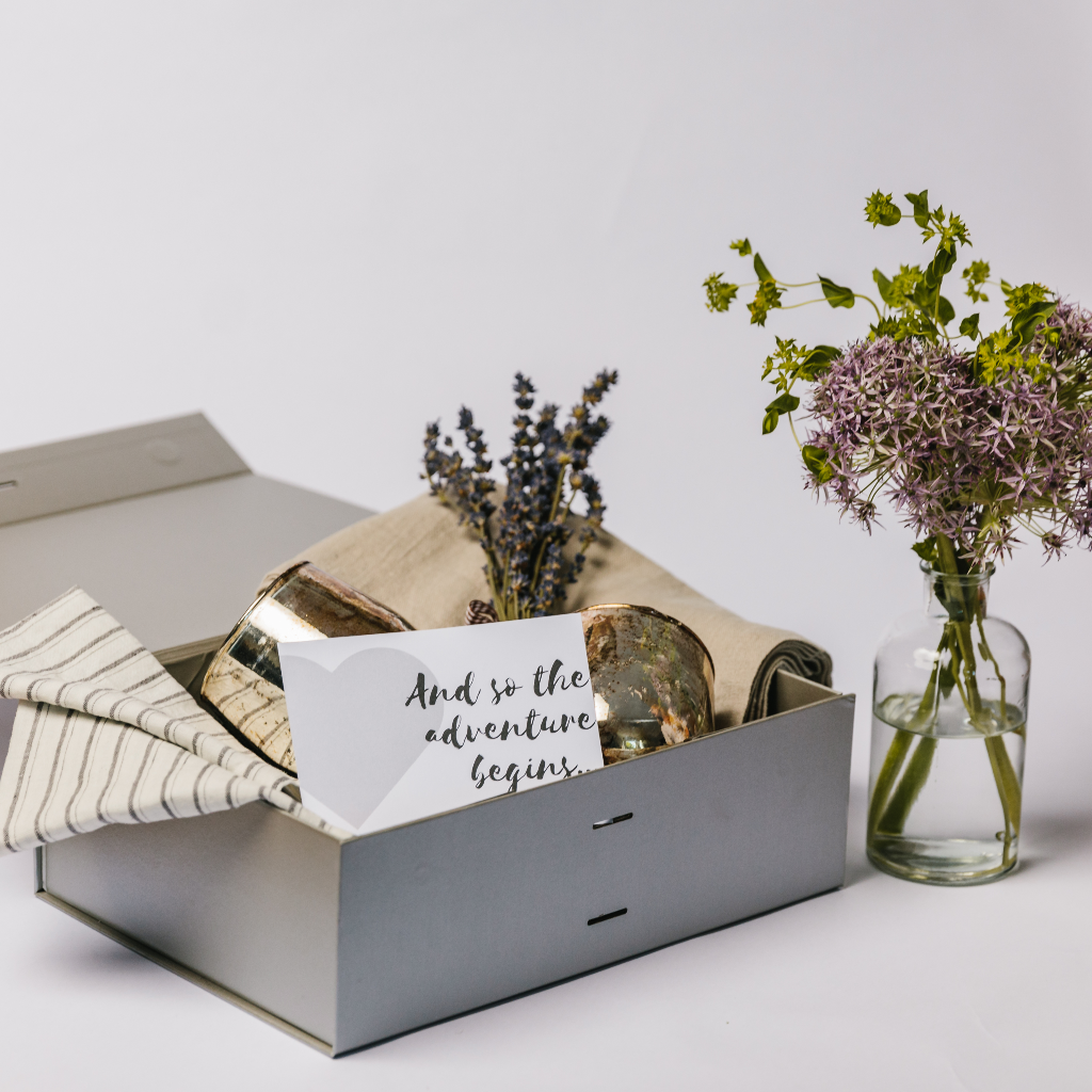 Image of full 'The Adventure Begins' gift box containing bronze candle holders, linen table napkins and runner, a vase with purple flowers and a posy of dried lavender