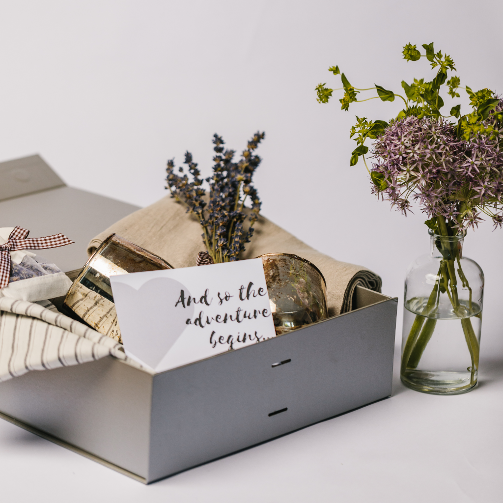 Tableware within luxury silver gift box laid out to show a bud bottlevase, bronze candle holders, a posy of dried lavender, striped linen napkins and a natural coloured linen table runner