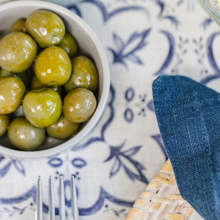 Top down view of green olives in a small grey dish set on a linen runner with blue and white mosaic pattern.