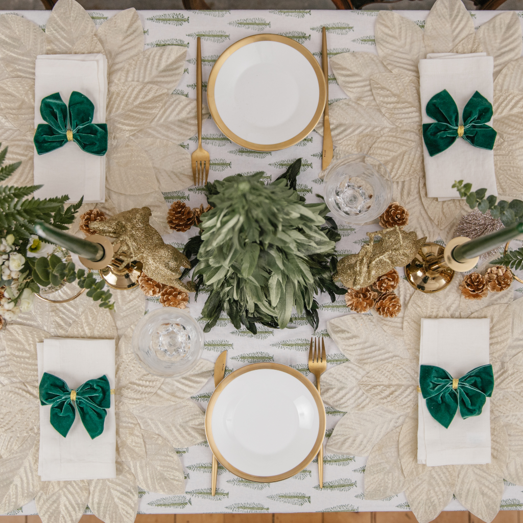 Top down view of four person Christmas tablescape including green feather trees, gold cutlery and golden pine cones scattered down table centre