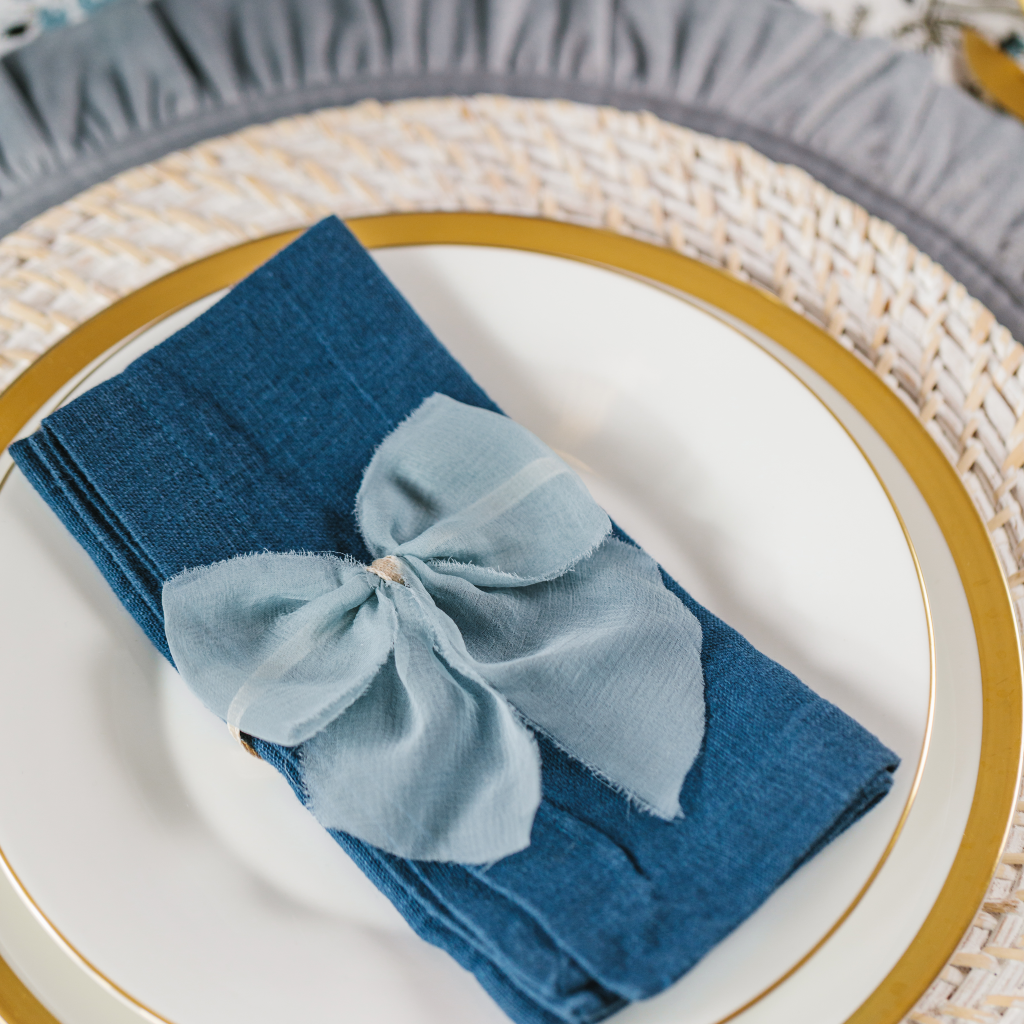 100% linen navy blue napkin folded and tied with pale blue raw silk napkin bow and set on a stack of white and gold plates.