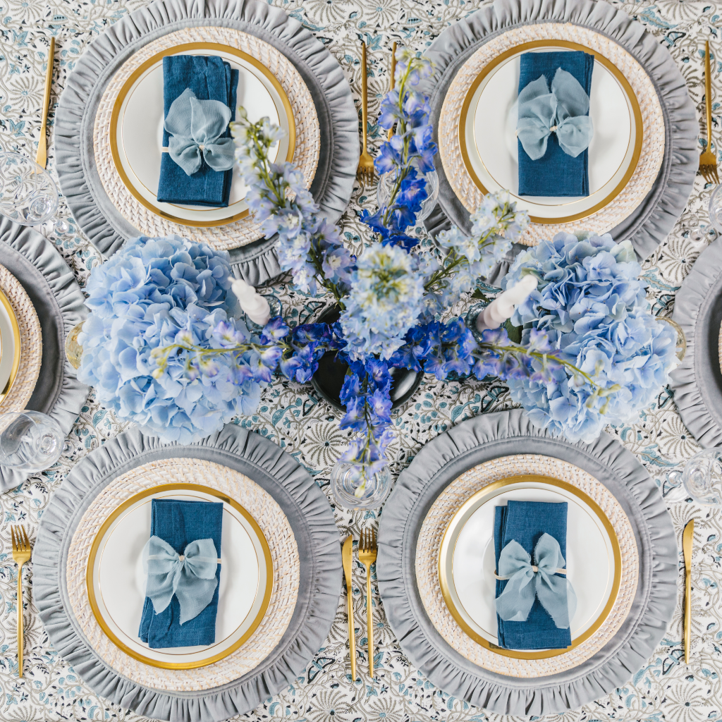 Top down view of Truffle's blue Twilight Collection including large blue flower central display and white and grey twist candles. The four place settings include mink grey velvet ruffled placemats, white wash rattan charger plates, navy linen napkins and Indian block print table cloth.