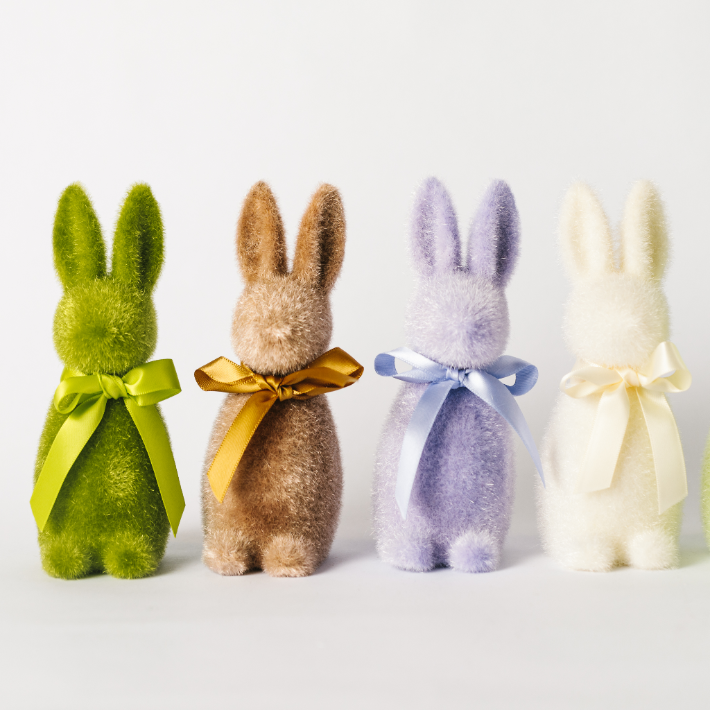 Mini Easter bunny decorations lined up in a row - moss green, soft brown, lilac and cream flocked statuettes with ribbon ties round the neck.
