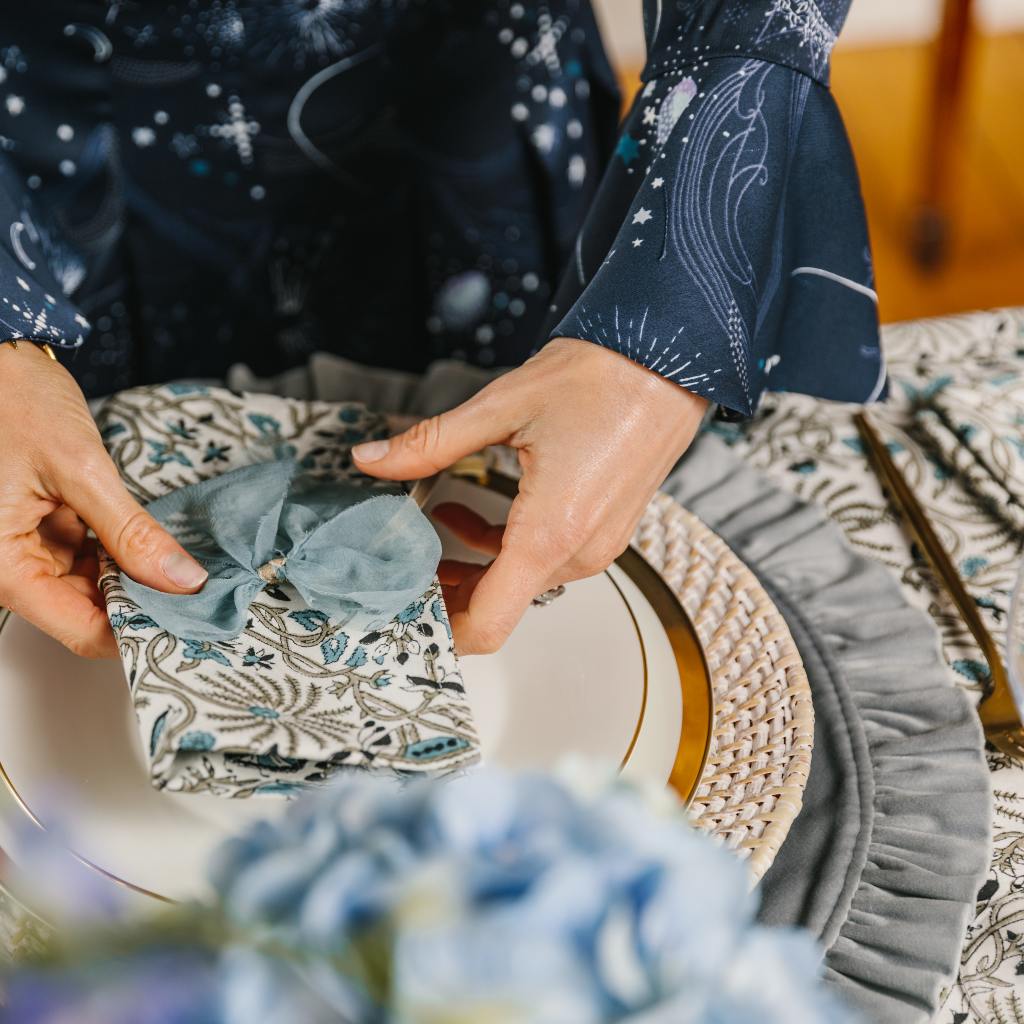 Hands tying  a blue and white Indian block printed napkin with a pale blue raw silk napkin bow. The place setting includes a white wash rattan charger plate and mink grey ruffed velvet placemat.