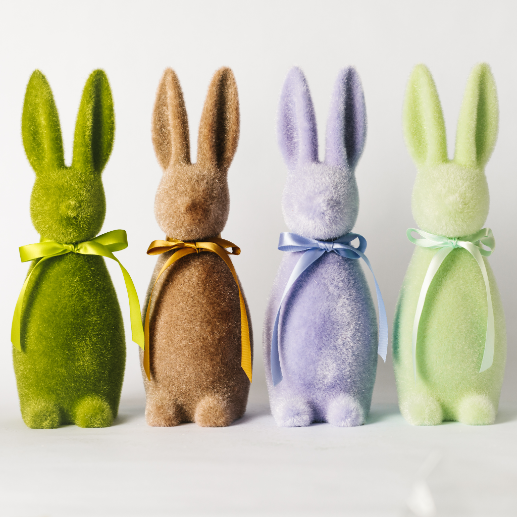 Row of large flocked Easter bunnies in moss green, soft brown, lilac and mint green. The bunny statuettes are all tied with a ribbon bow tie and set at 30cm tall.