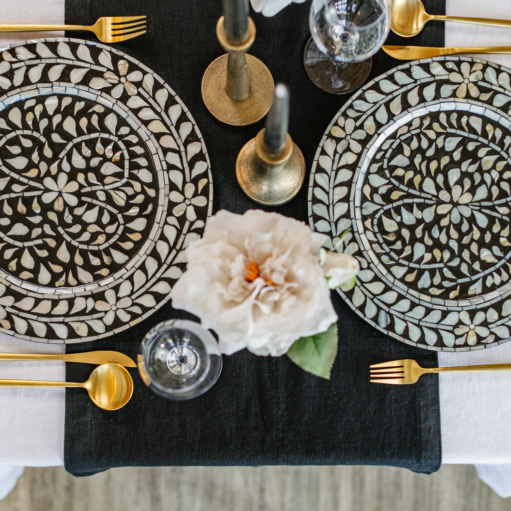 Top down flat lay image of black linen table runner, statement black and white mother of pearl inlay charge plates, brass candlesticks, black tapered candles and white rose blooms